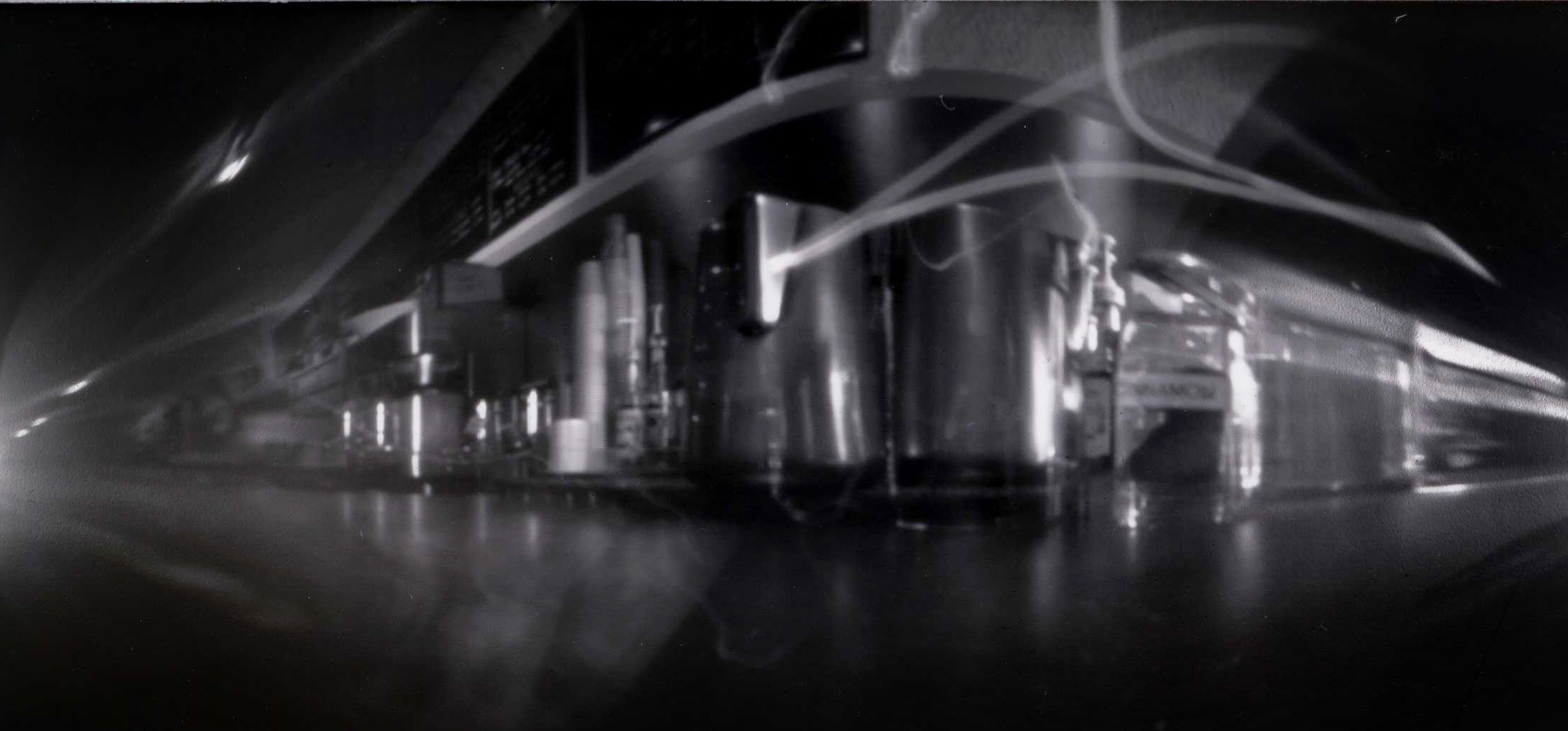a black and white pinhole camera photograph with a large amount of curvature distortion except at the center of the image; this is a photograph of the stainless steel milk pitchers sitting on a barista’s counter in a coffeeshop; most of the image is too distorted to discern except for the milk pitchers but there are stacks of cups, the espresso machine, and the menu boards to the left and a shakers of cinnamon to the right; there are trails of light showing that during the exposure of the image, the milk pitchers were moved by the barista